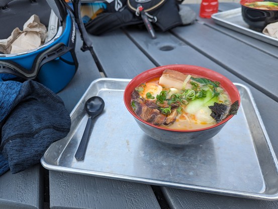 A photo of a bowl of ramen on a metal tray. There is a spoon on the side of the tray and my helmet can be seen on the table.