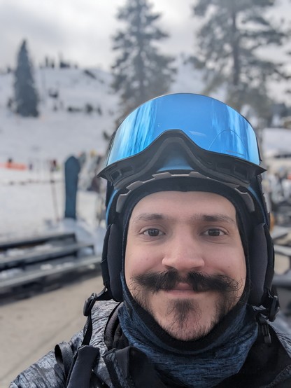 A selfie with my helmet and goggles. Behind me are trees and ski runs.