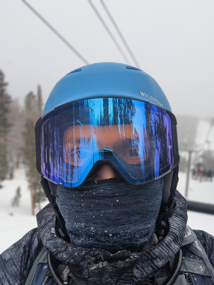A selfie while sitting on a chair lift. I am wearing my helmet, goggles, and a balaclava and all you can really see are my eyes through the goggles. Behind me are trees and lots of snow.